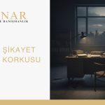 Gece vakti loş ışıkta bir hukuk bürosu masası; üzerinde belgeler, dosyalar ve bir masa lambası bulunuyor. Arka planda şehir manzarası ve gölgede beliren bir figür, dava veya şikayet edilme korkusunu simgeliyor.